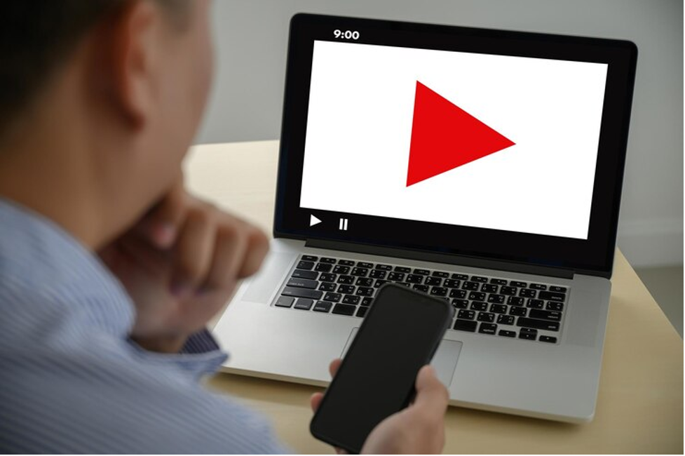 Seorang pria sedang menonton video di laptop sambil memegang smartphone, sebagai ilustrasi cara mendownload video dari YouTube.