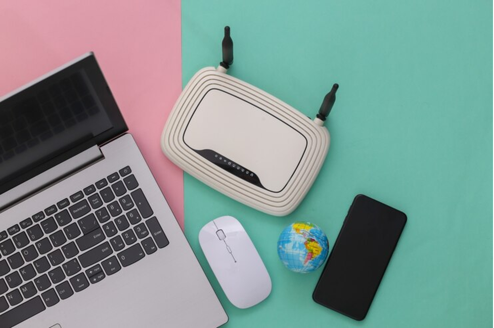 A silver laptop beside a white router, a wireless mouse, and a smartphone, all placed on a pink and green background.