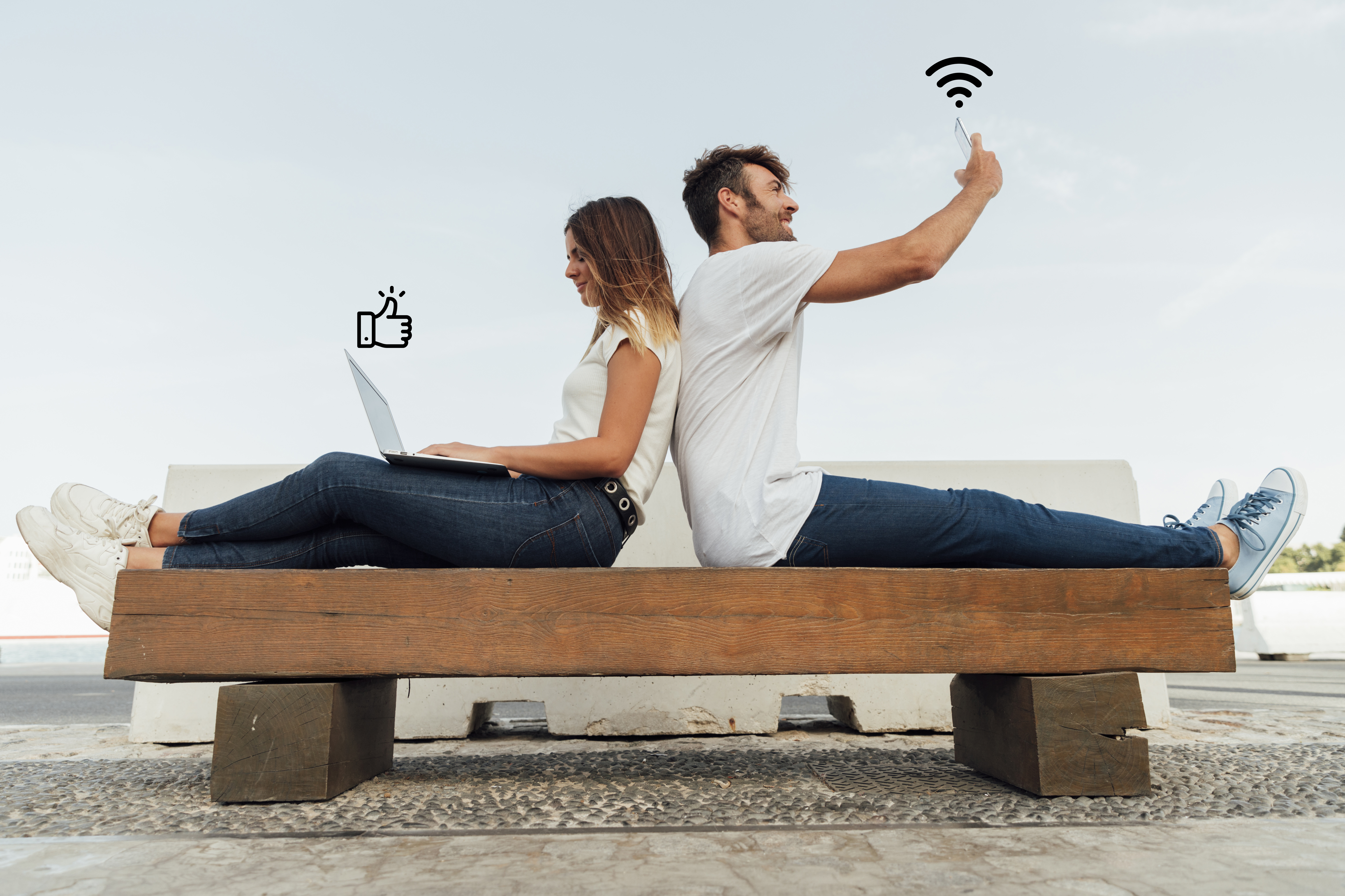 A couple sitting on a bench, one holding a laptop and the other holding a phone, illustrating cara berbagi sandi WiFi.