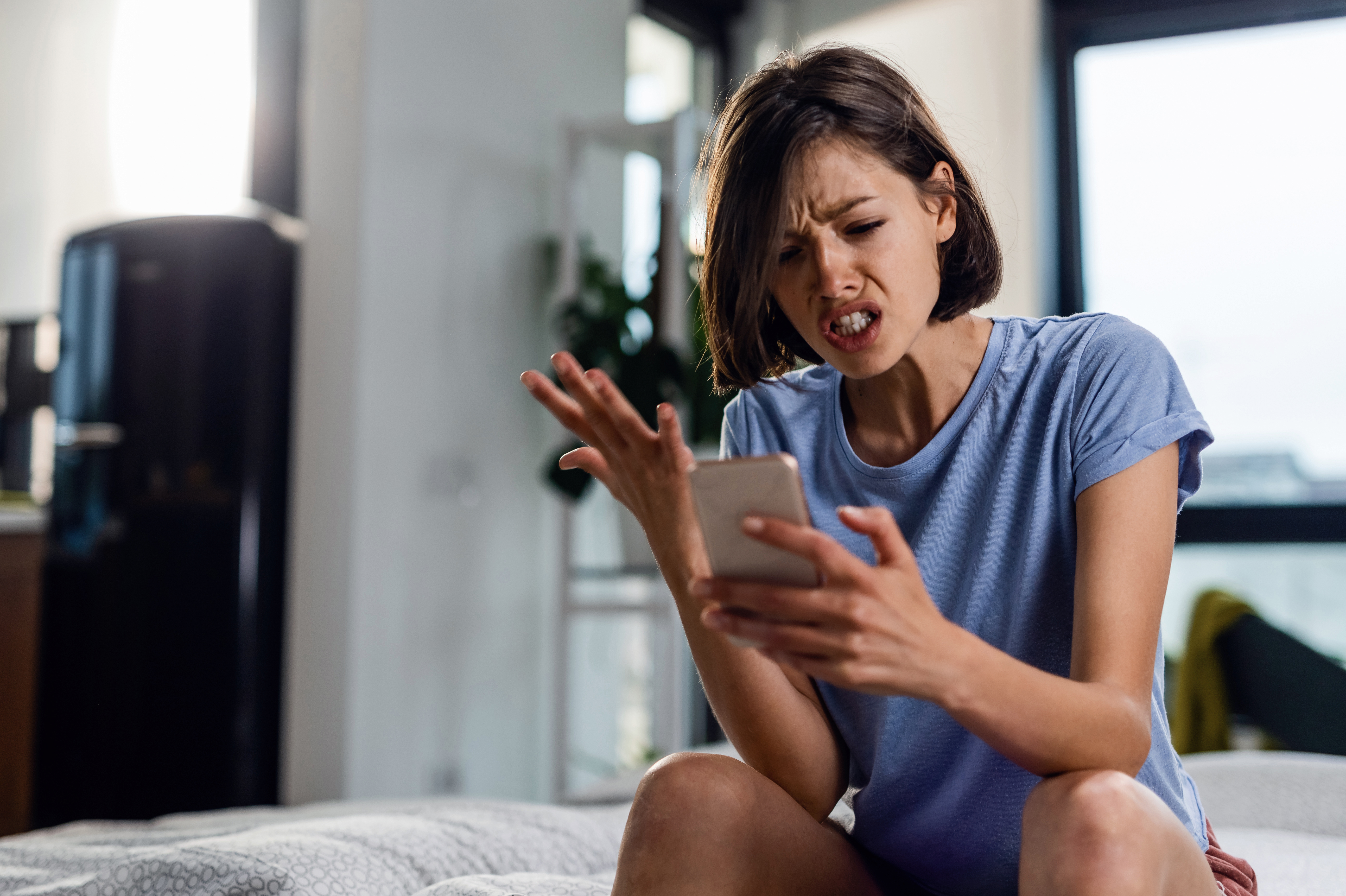 Young frustrated woman reading text message cell phone in disbelief while sitting on the bed at home.