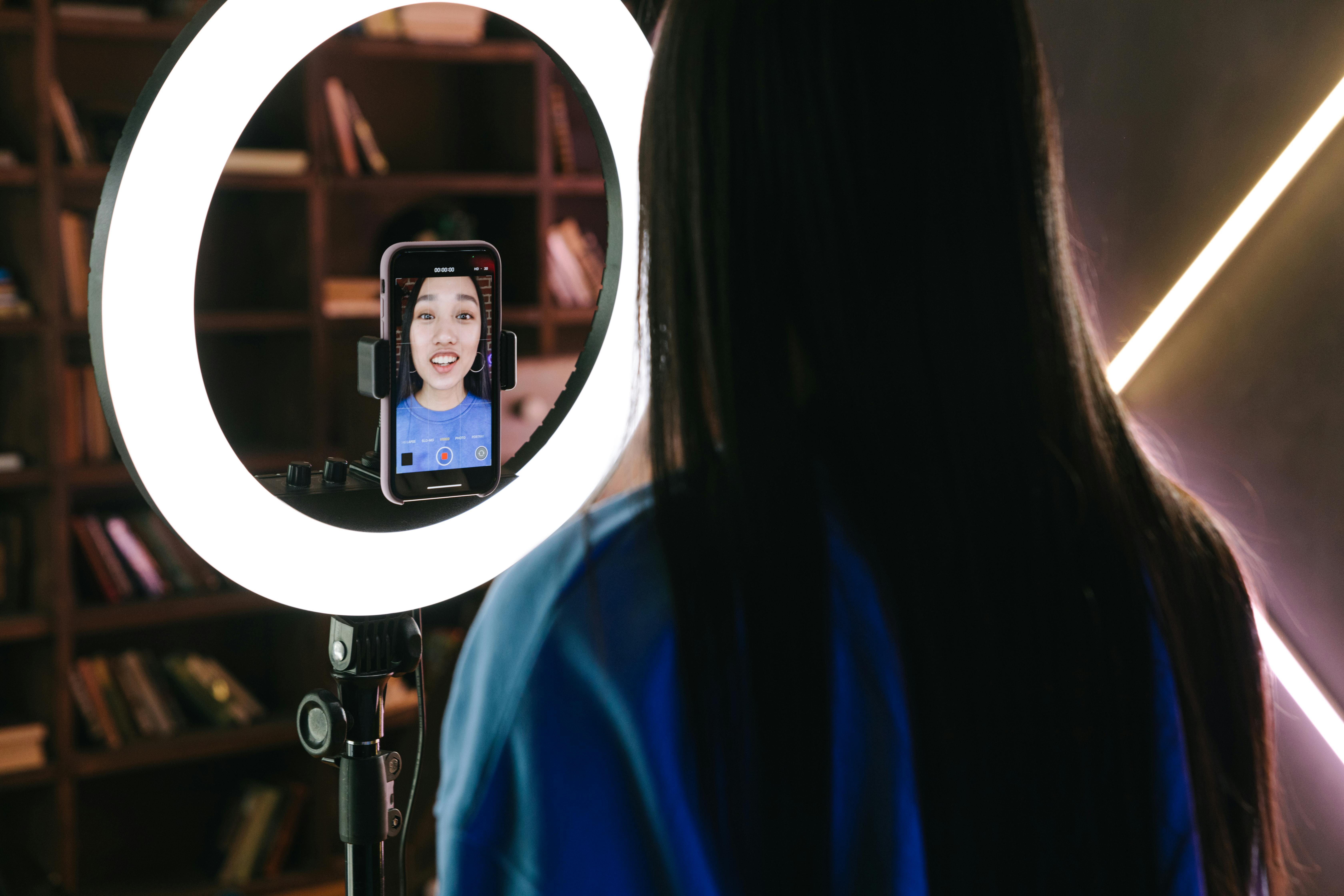 Woman recording herself with a smartphone mounted on a ring light tripod, illustrating how to go live on TikTok (cara live di TikTok).