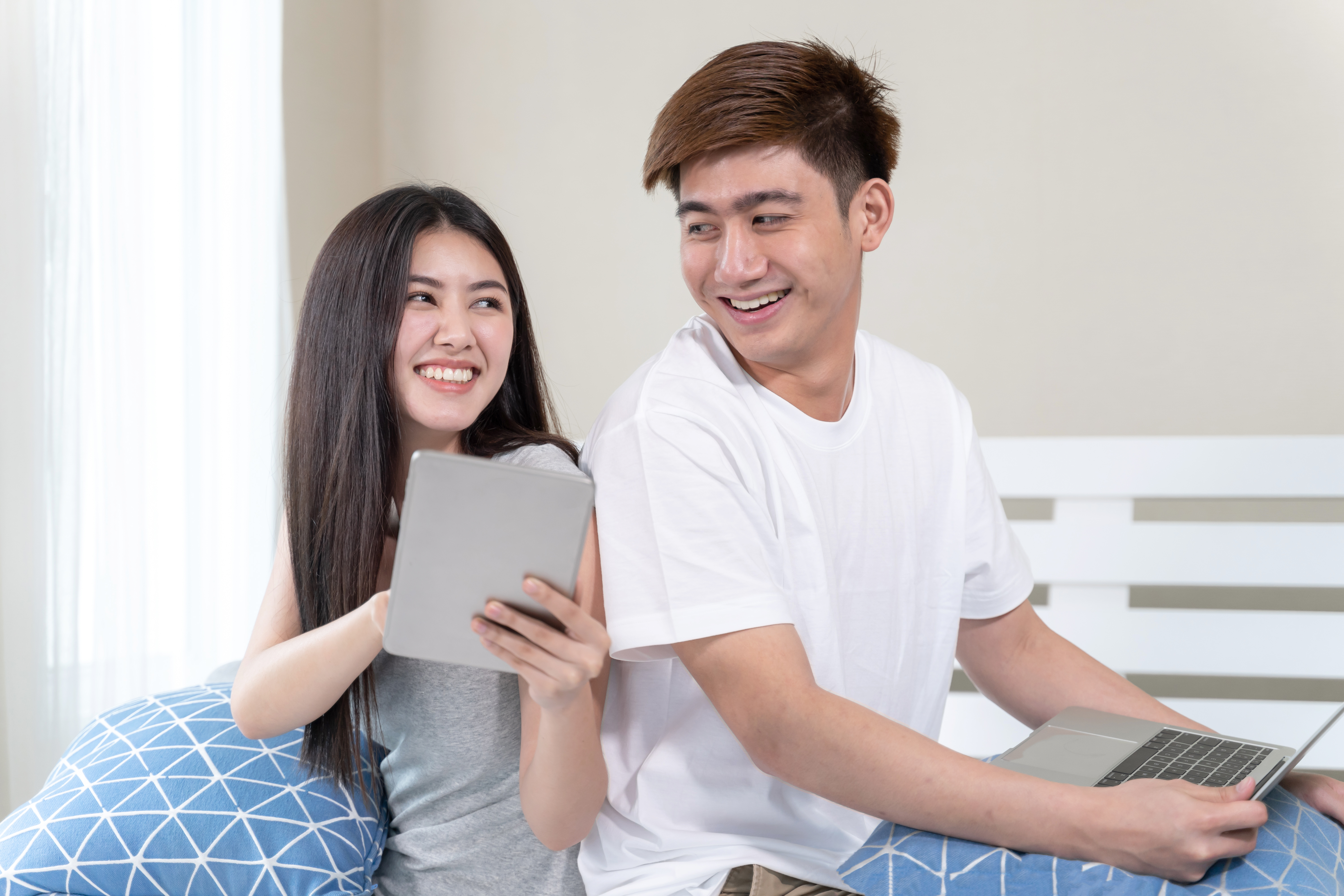 Young woman happily chatting on smartphone and man using laptop with joy while sitting together on a bed, illustrating shared internet access using a WiFi dongle at home.