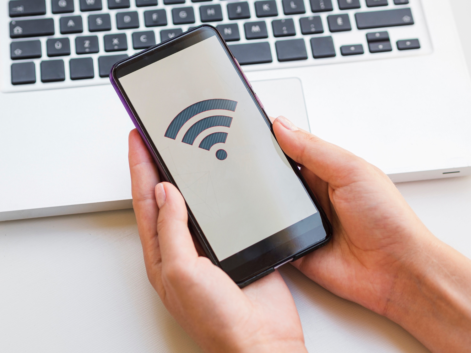 Close-up of a hand holding a smartphone in front of a laptop, illustrating reliable WiFi connection in Bandung (WiFi terbaik Bandung).