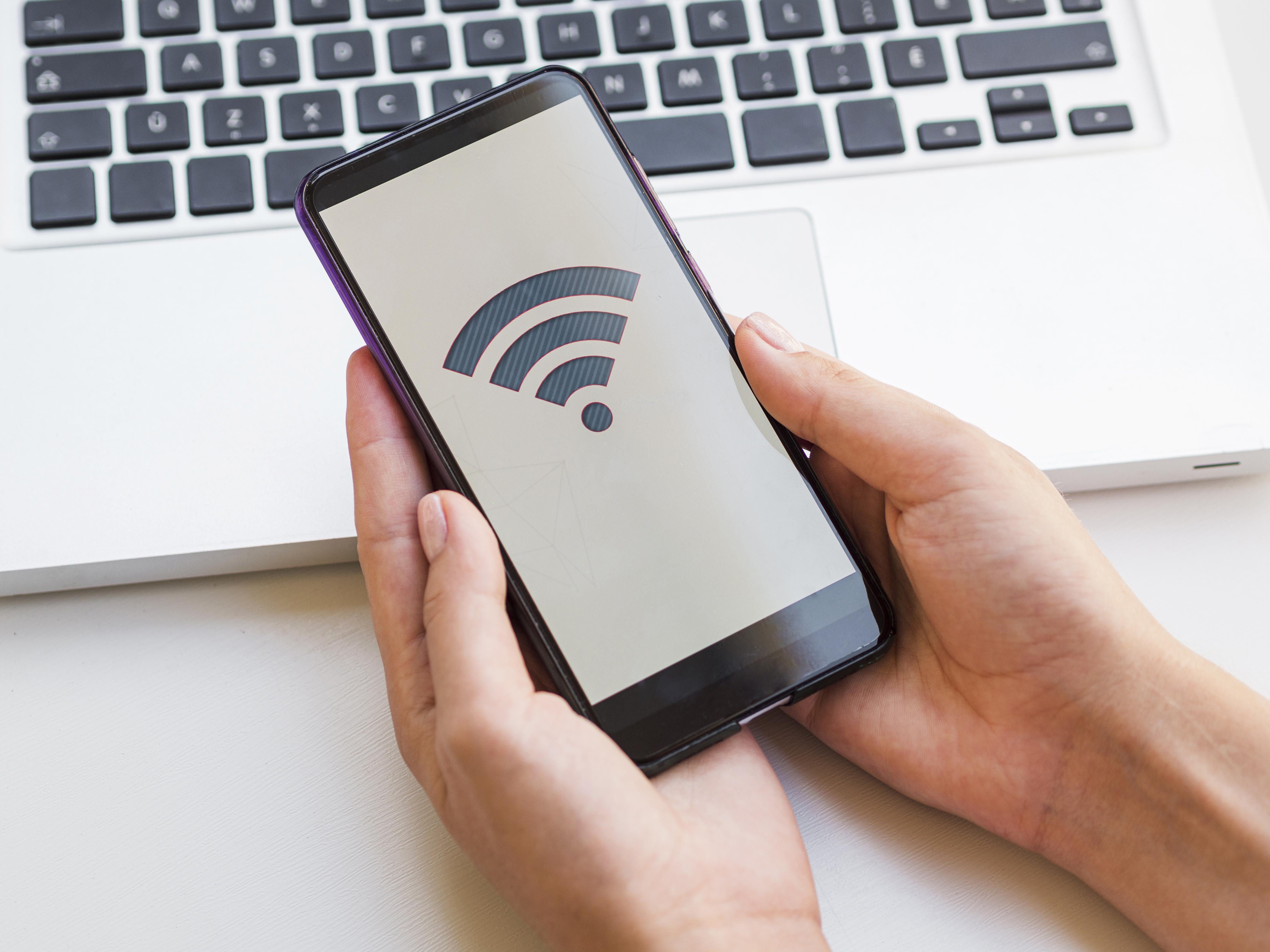 Close-up of a hand holding a smartphone in front of a laptop, representing access to high-speed internet in Indonesia (provider internet terbaik Indonesia).