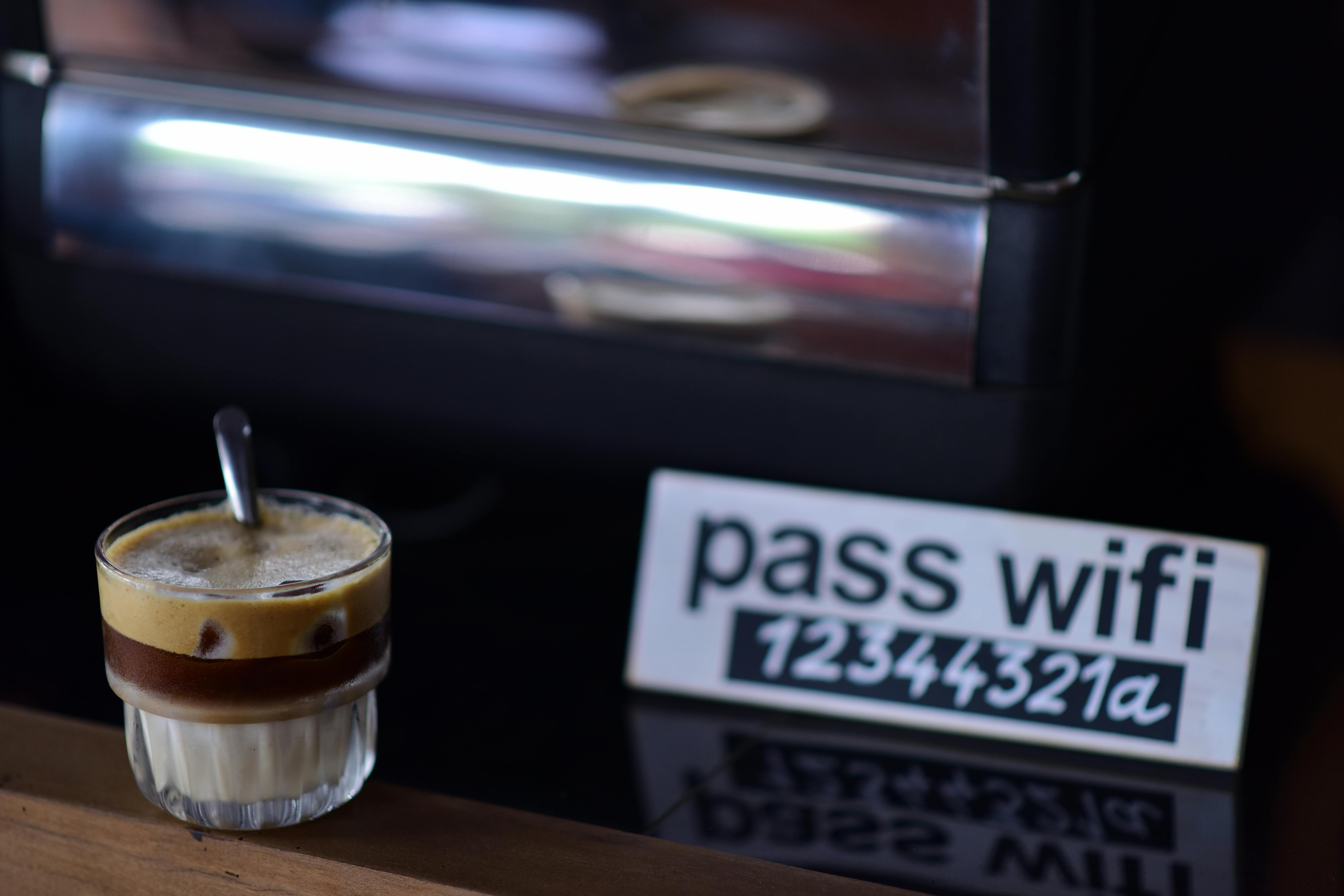 Glass of espresso coffee in a cozy café setting, with a note beside it that says 'pass wifi 12344321a', illustrating a casual reminder for how to change WiFi password (cara ganti password wifi).