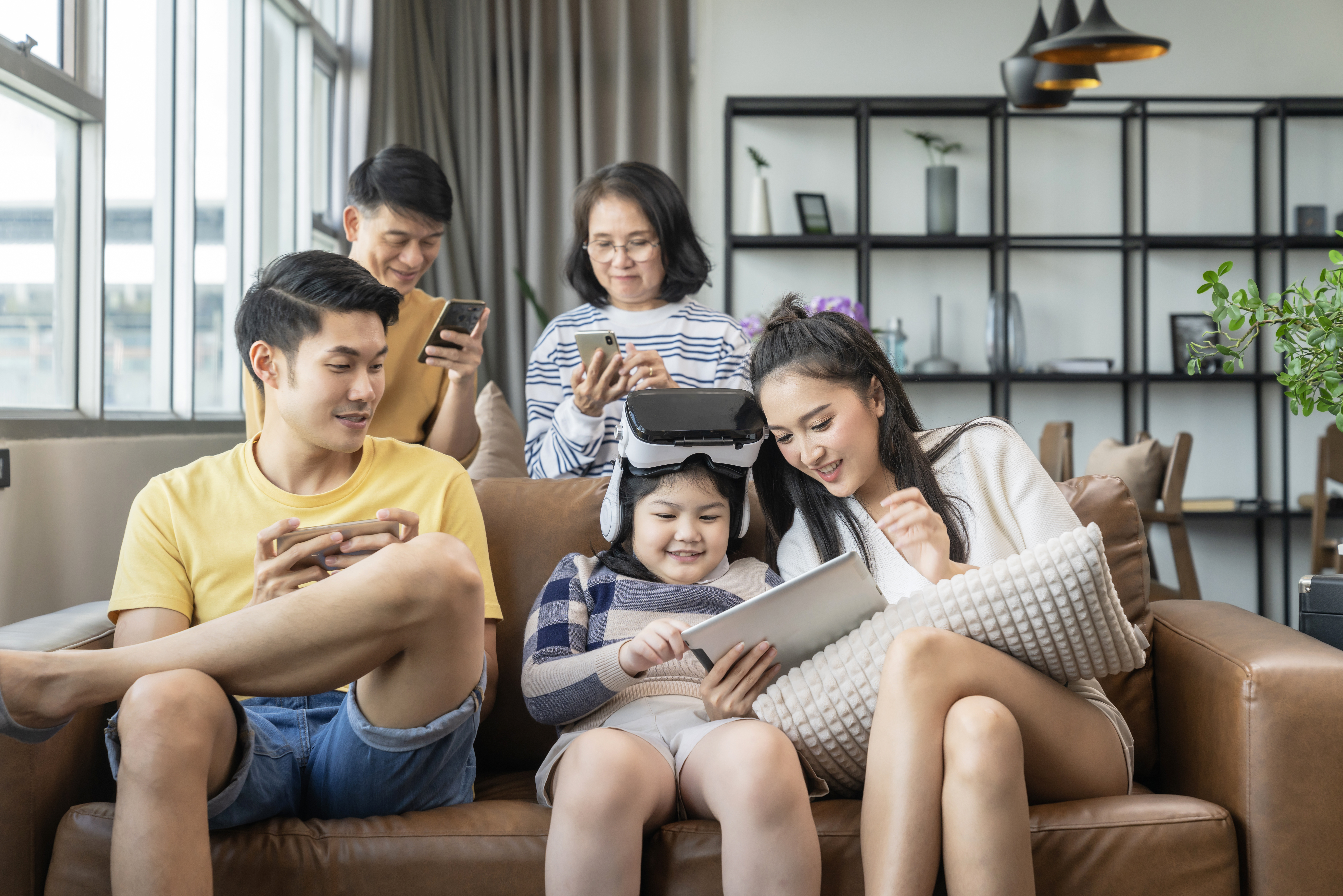 Asian family enjoying smart devices like smartphones, tablets, and VR at home, showing the benefits of installing WiFi (pasang WiFi) for the whole household.