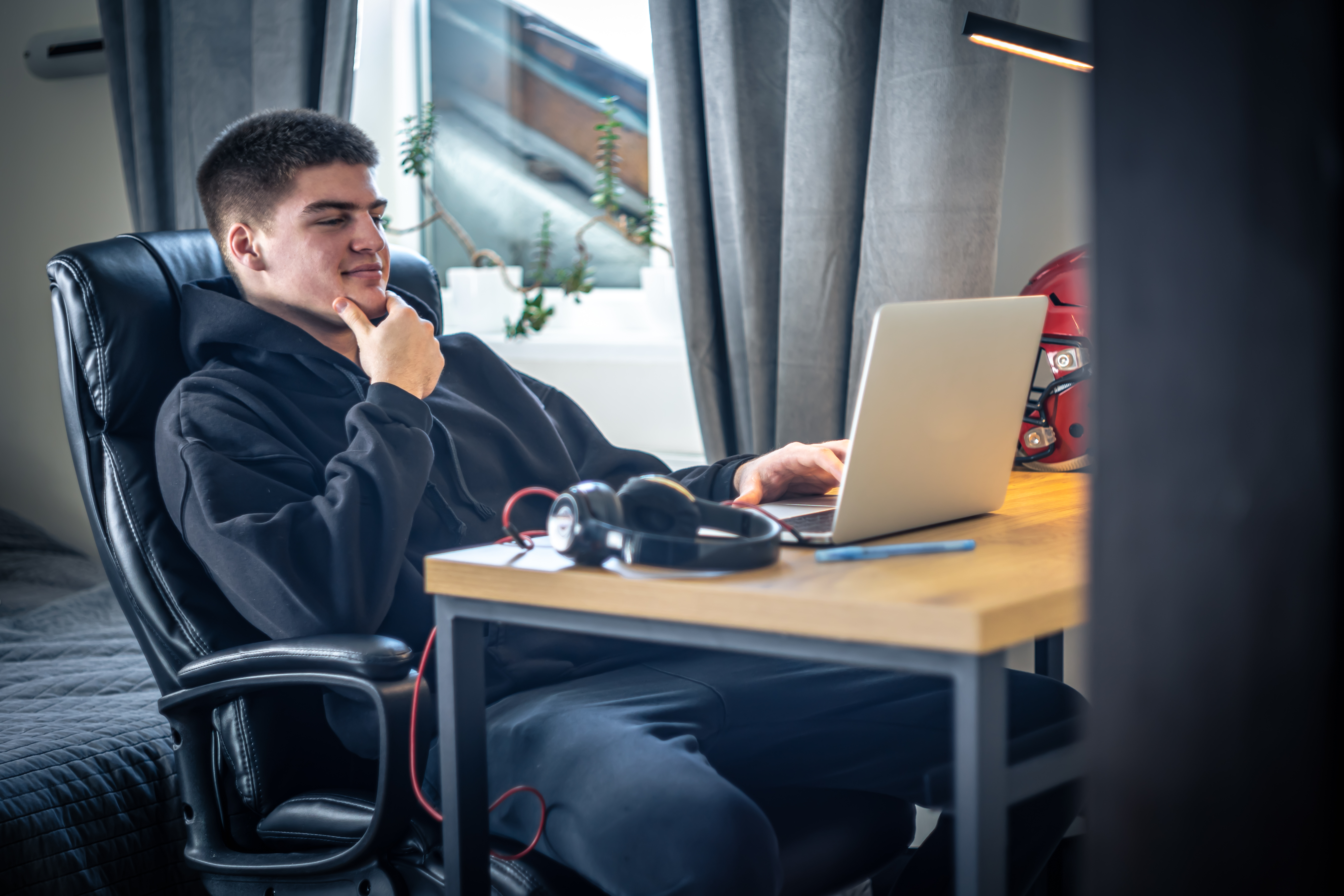 A man wearing headphones sitting at a laptop in his room, sports helmet visible in the interior, to illustrate light PC games (game PC ringan)