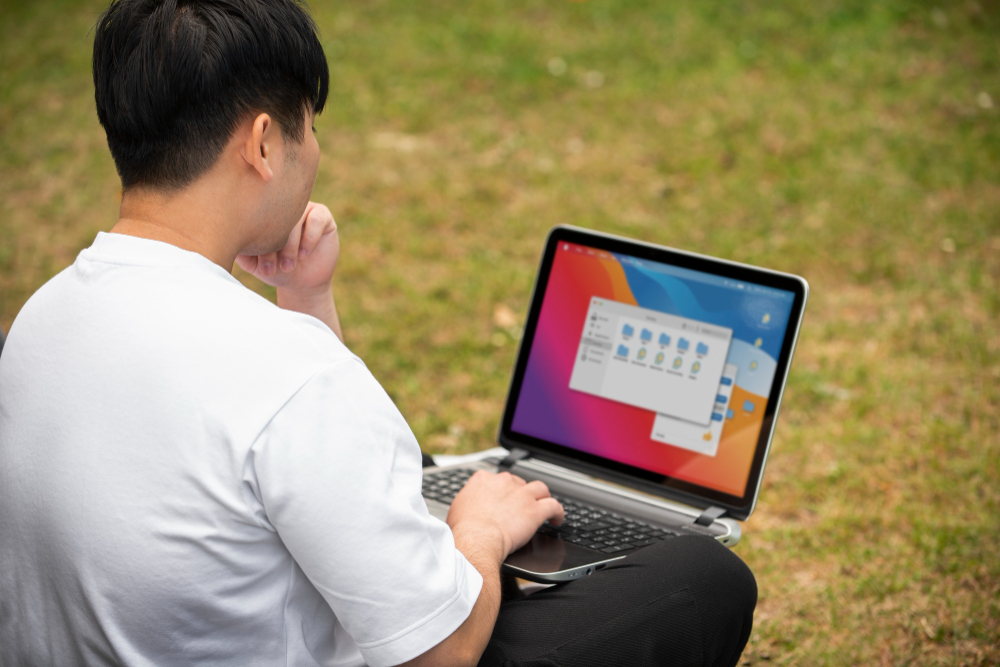 A male college student studying outdoors with a laptop on his lap, representing someone searching for or practicing how to record on a laptop (cara screen record di laptop).
