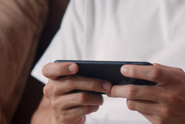Young Asian man lying on a sofa in the living room, smiling while playing a strategy game (game strategi) on his smartphone during leisure time.