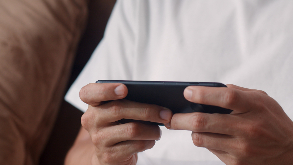 Young Asian man lying on a sofa in the living room, smiling while playing a strategy game (game strategi) on his smartphone during leisure time.